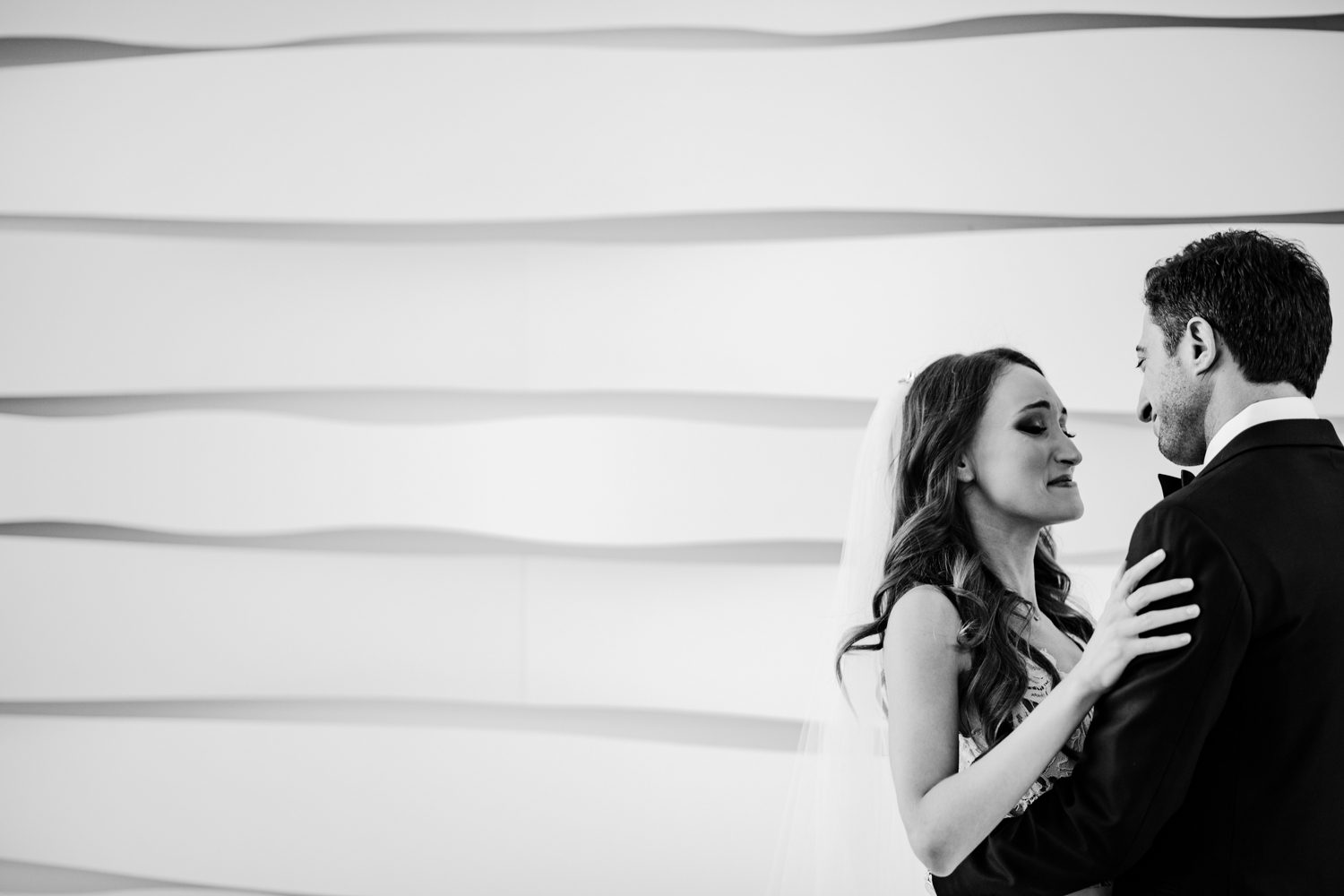 A bride and groom embracing in front of a white wall.