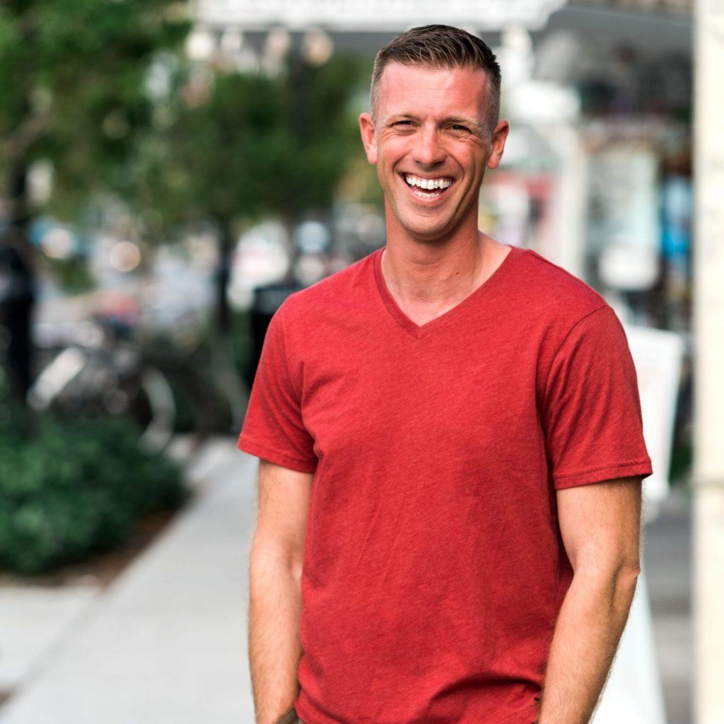 A smiling man in a red v-neck shirt standing on a sidewalk, captured by a Key West Wedding Photographer.