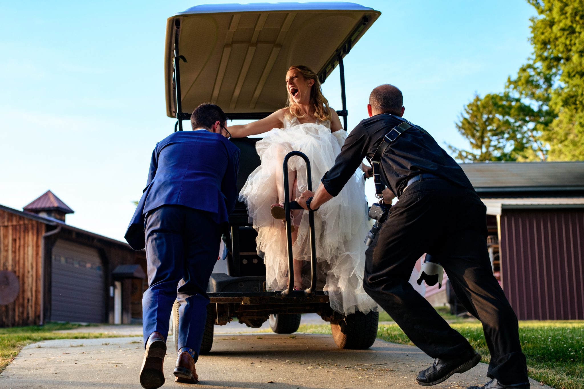 Groom and wedding photographer pushing a stalled golf cart on a sunny day at an The Farm Events wedding, while a laughing bride watches from the passenger seat, captured by a second photographer in Candler NC