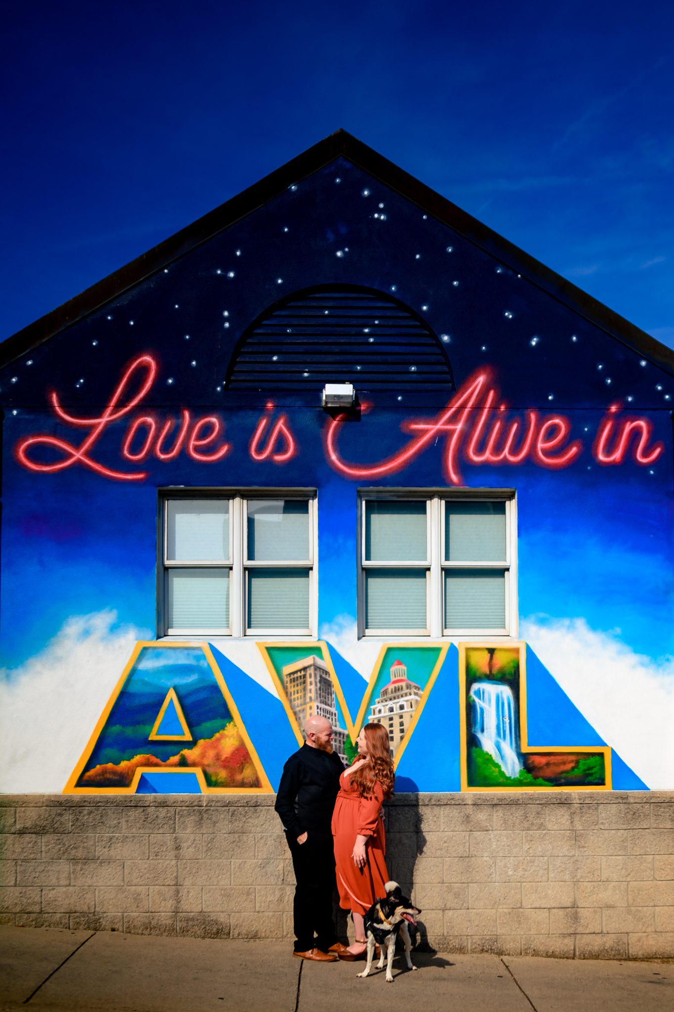 engaged couple standing with their dog in front of the love is alive in asheville mural on the side of spicer greene jewelers in downtown asheville