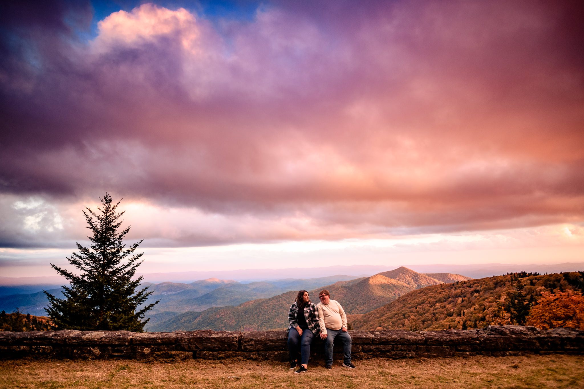 engaged couple sitting on rock wall with pink sunset and mountains behind them