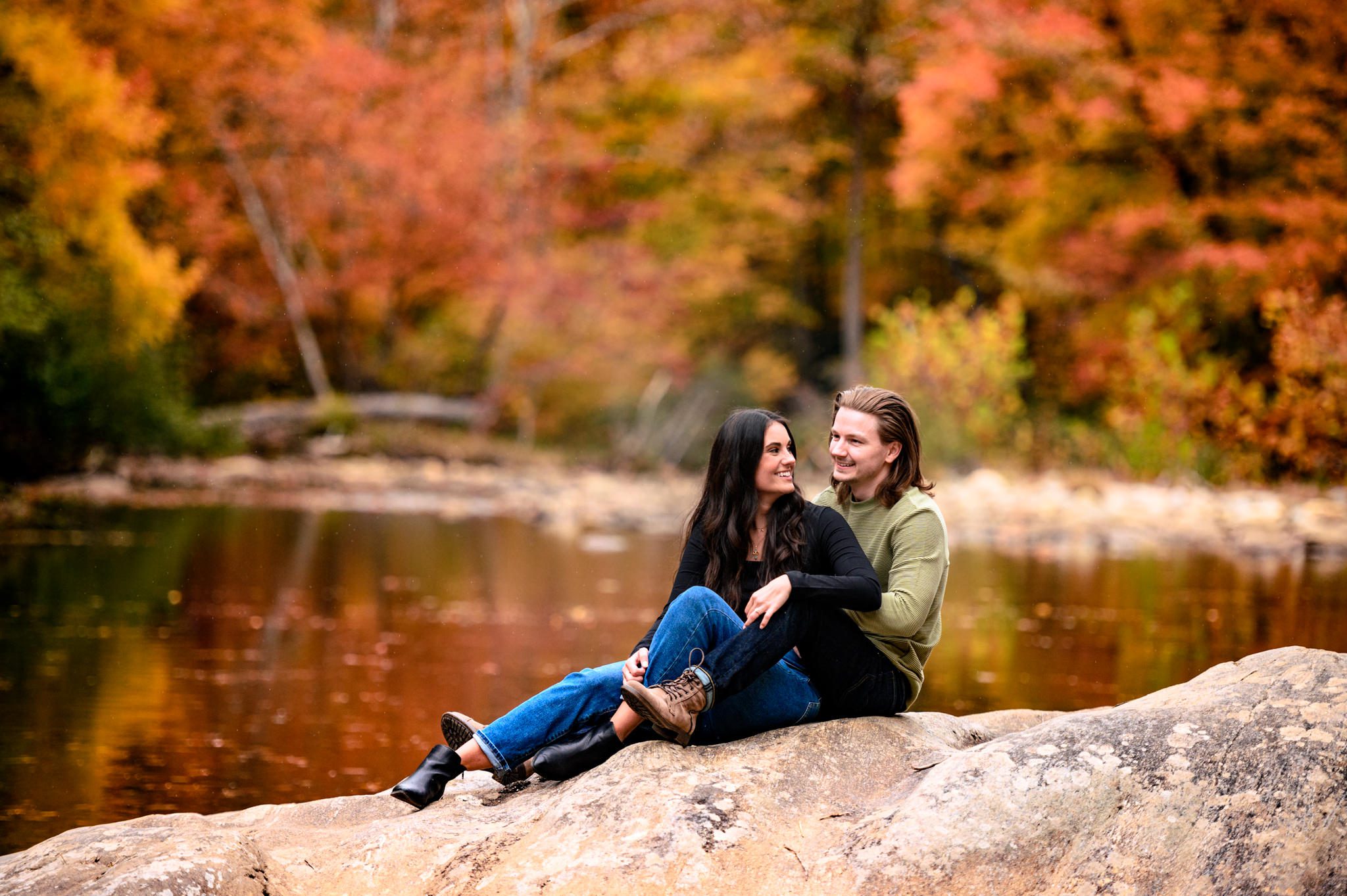 engaged couple sitting on rock in middle of river with fall colors behind them on the trees
