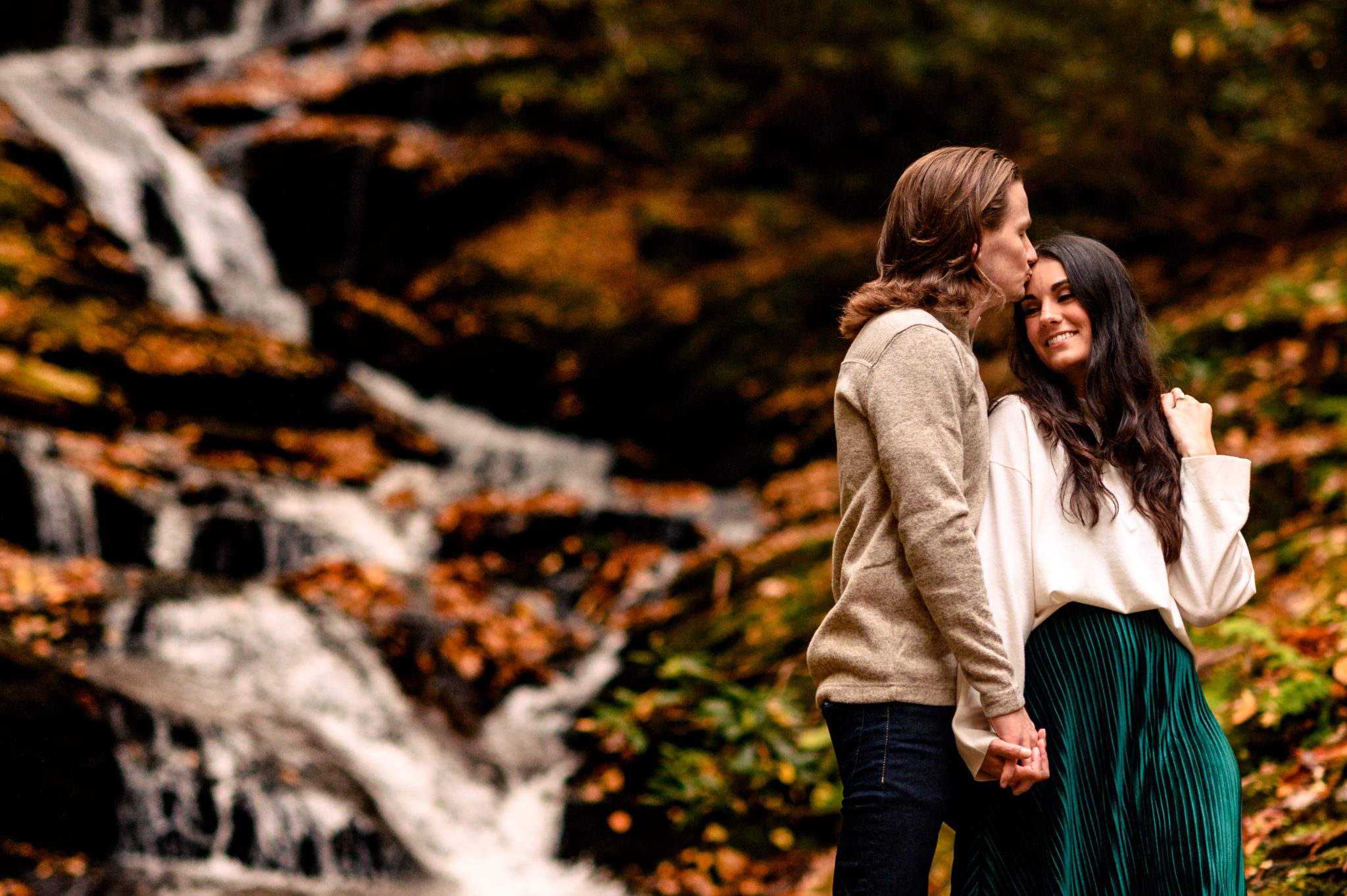 engaged couple smiling while groom kisses her forehead and waterfall behind them