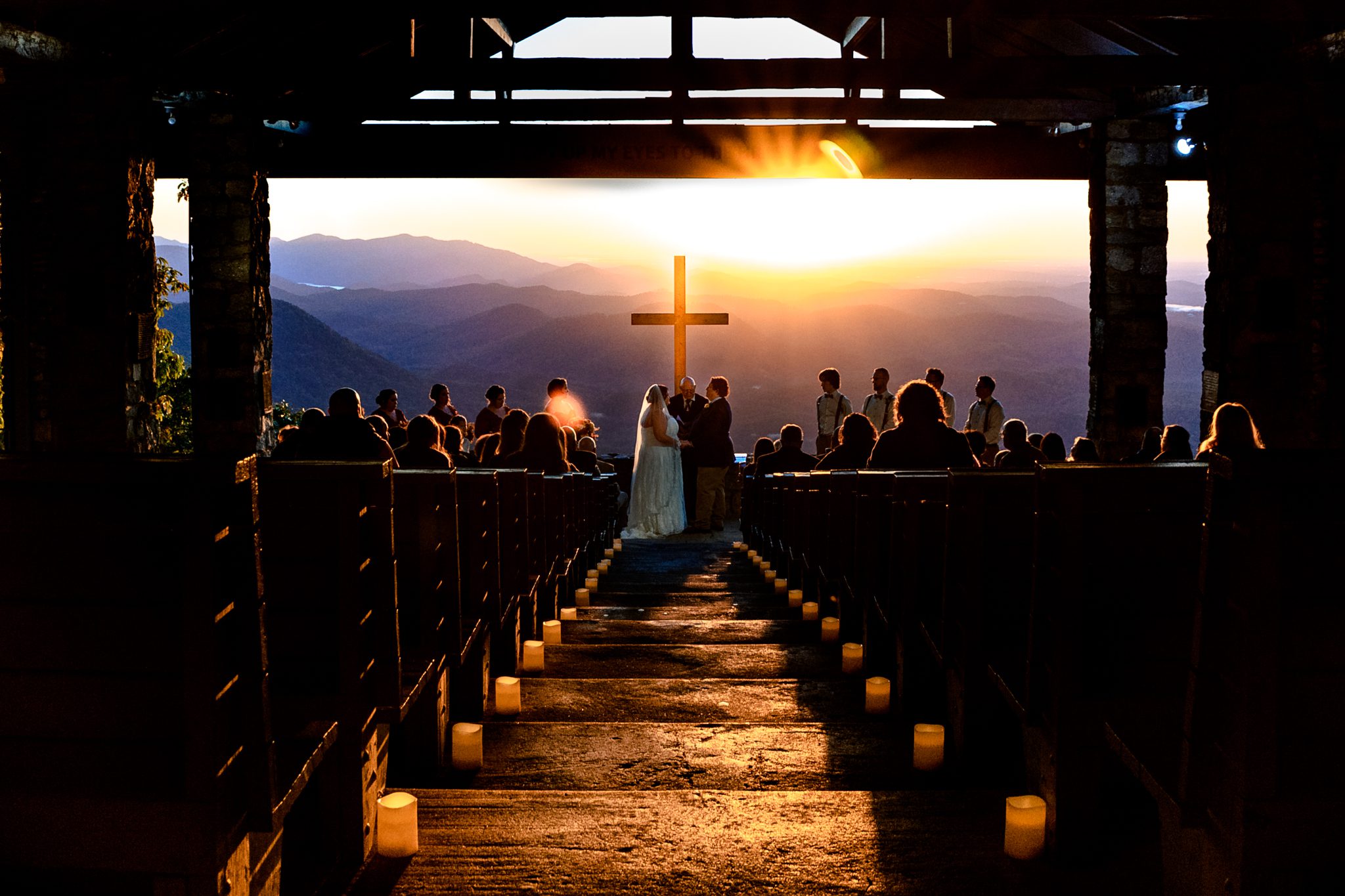 Bride and groom stand under illuminated cross with sunrise in the background at pretty place camp Greenville