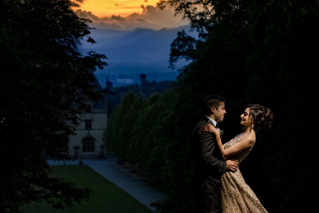 wedding couple taking sunset wedding photos overlooking the biltmore estate from the diana lawn. golden sunset over the mountains behind them