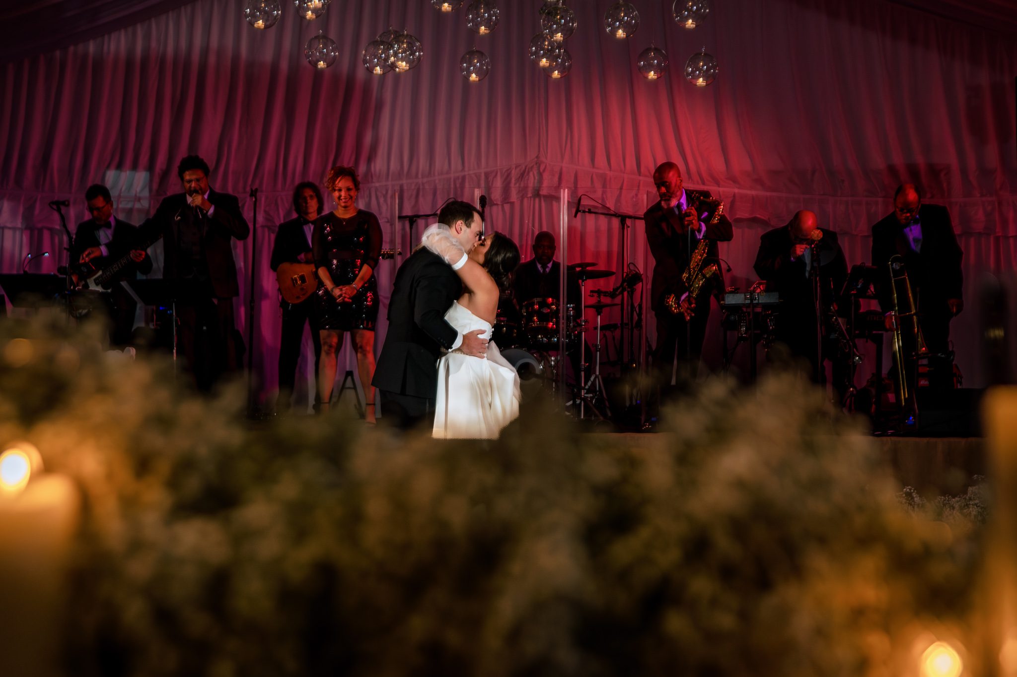 Bride and groom sharing the first dance and kissing with a band standing behind them on the stage with red lights