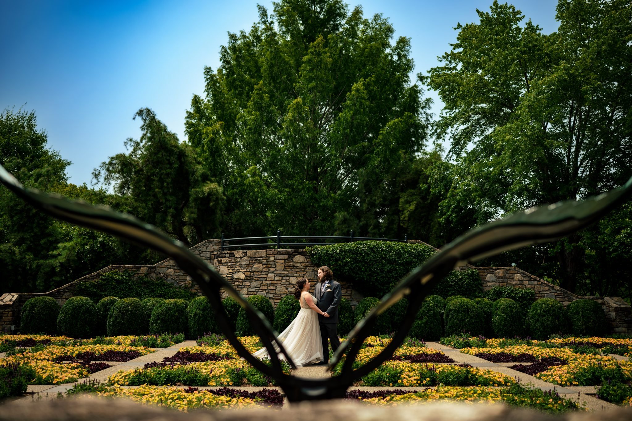 A bride and groom standing in front of a sculpture in the patch quilt garden at the NC Arboretum