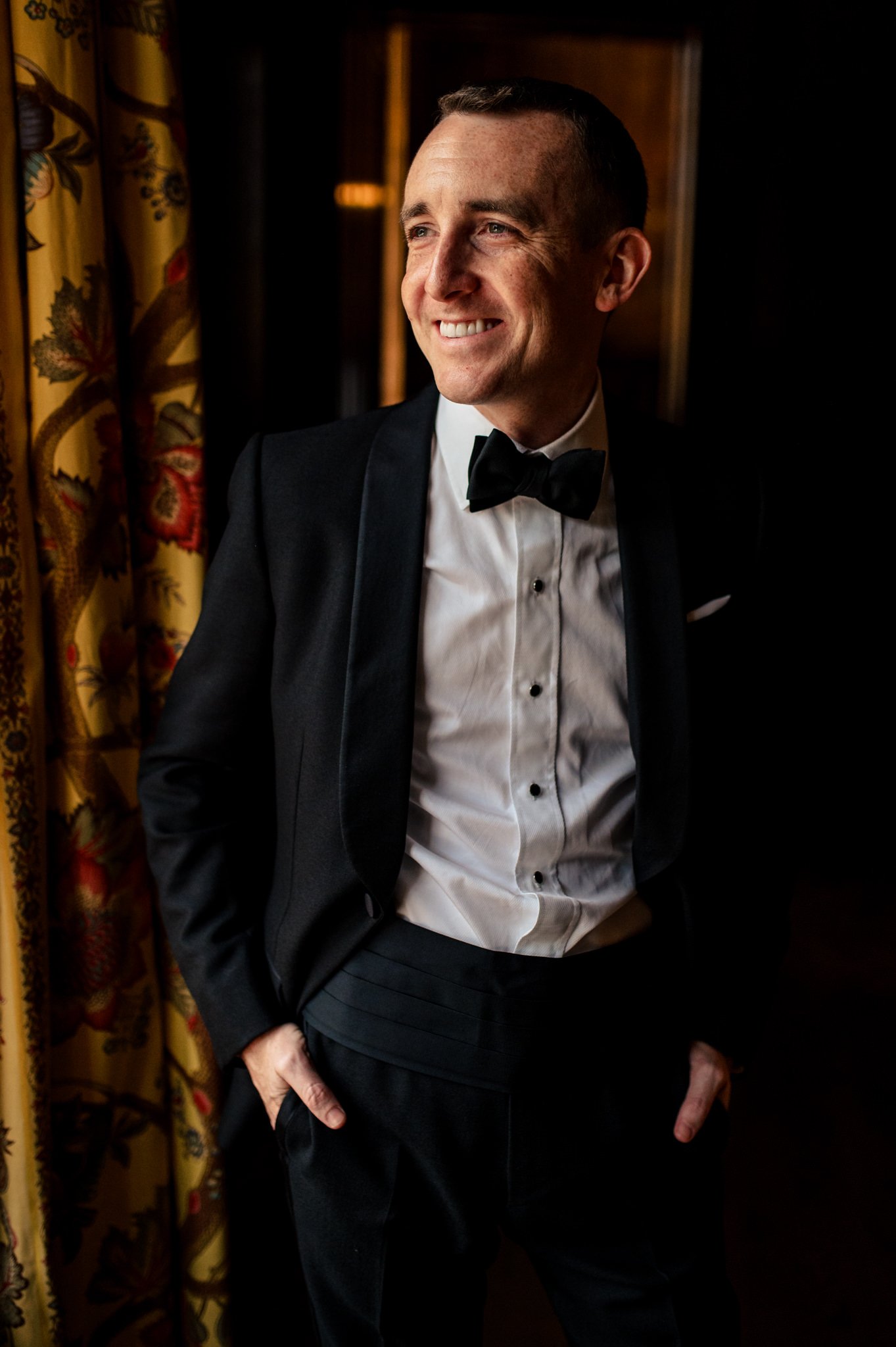 A man in a tuxedo smiles in front of a curtain.