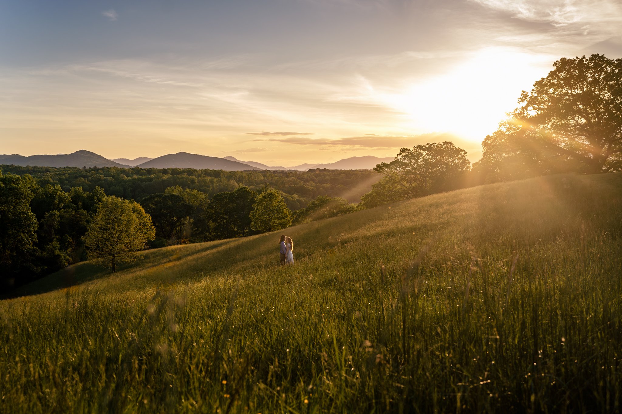 A couple walking in a grassy field on the Biltmore Estate at sunset with trees and distant mountains in the background.