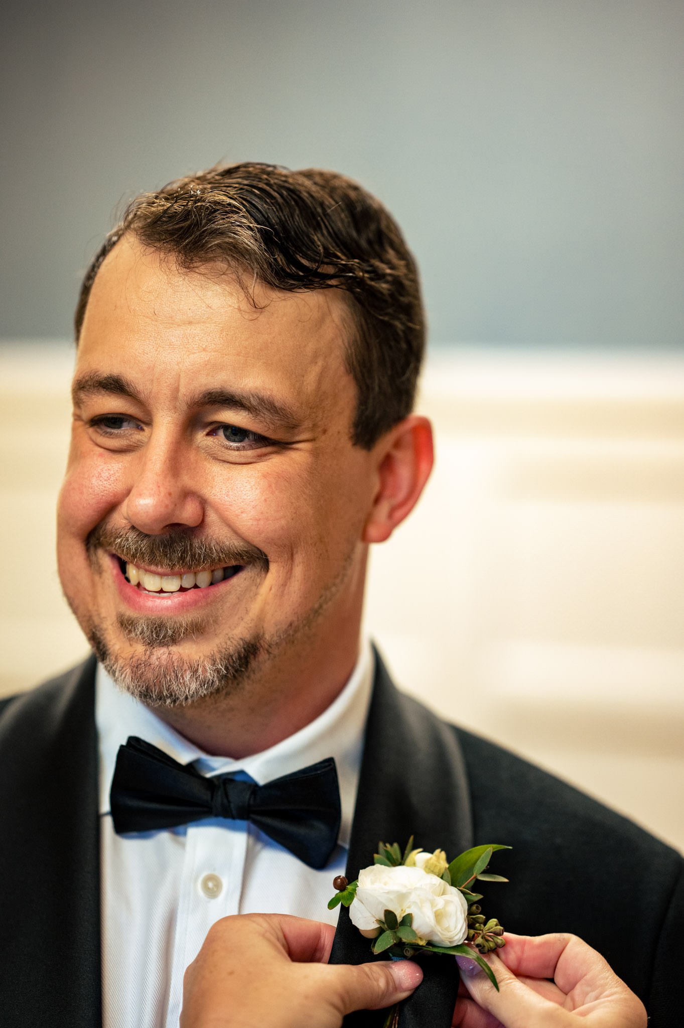 A smiling man in a tuxedo with a bow tie, adjusting a boutonniere on his lapel at a Homewood wedding in Asheville, NC.