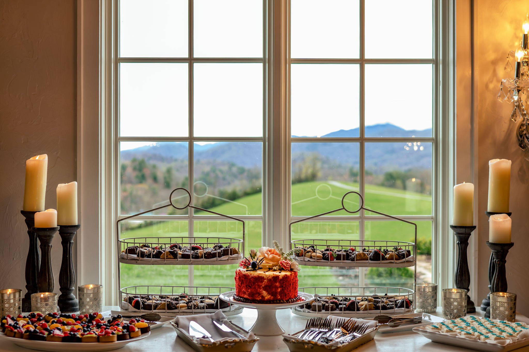 Elegant dessert table by a large window overlooking a scenic mountain view at The Ridge Asheville, featuring a cake, candles, and assorted sweets.