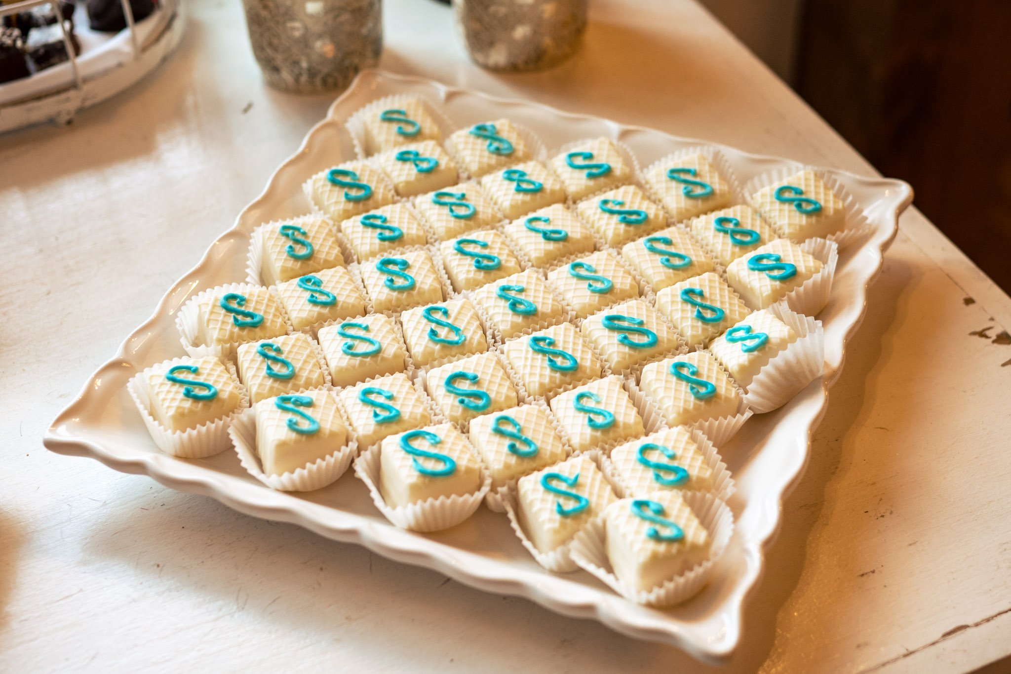 A tray of rectangular white chocolates decorated with a teal-colored letter 's' on top, presented on a white decorative plate at an Asheville wedding.