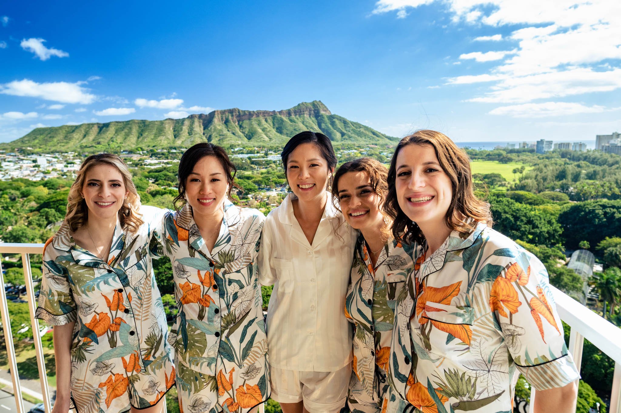 Five women smiling on a balcony with a scenic view of a lush green mountain and cityscape in Honolulu, Hawaii.
