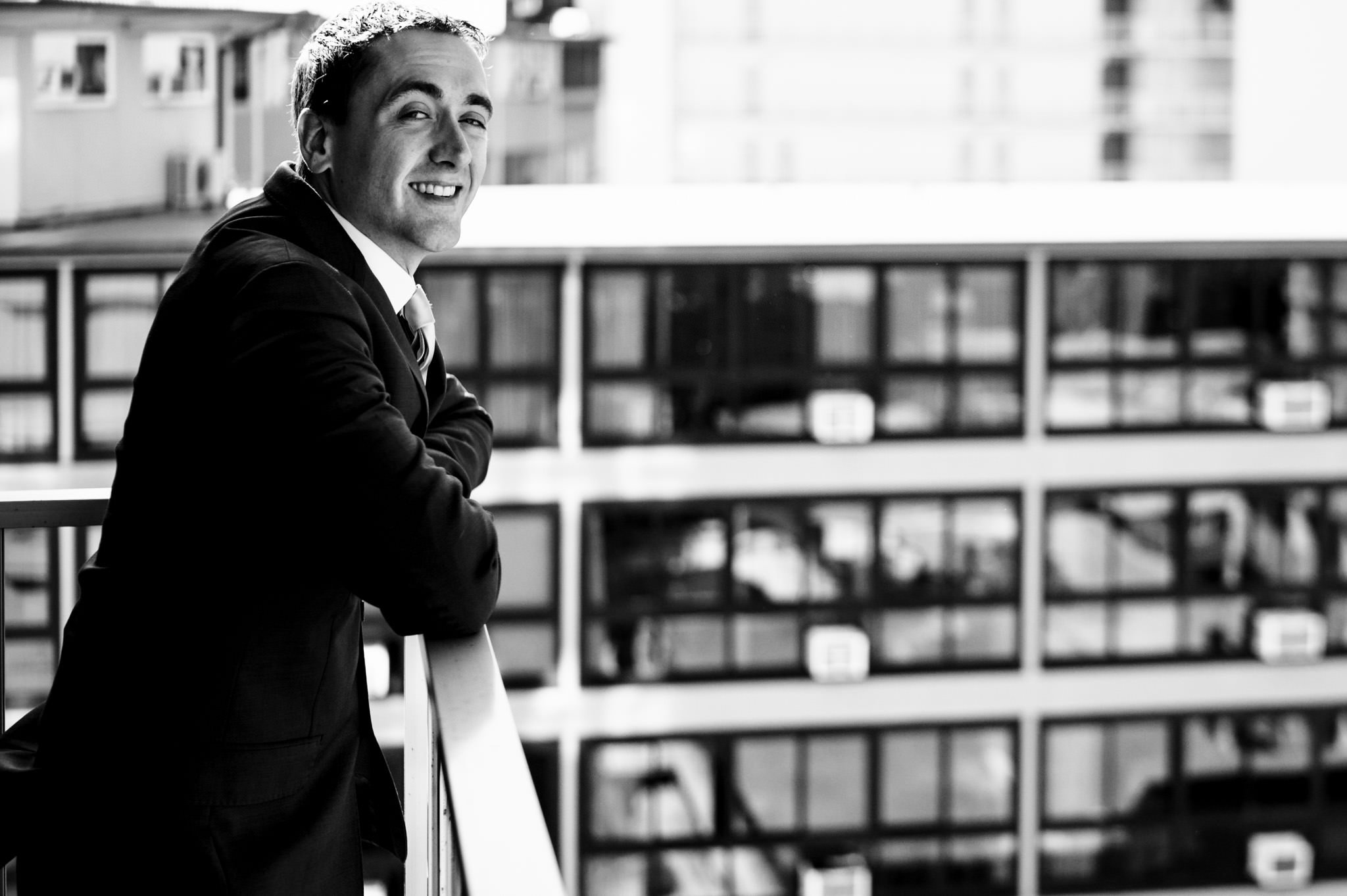 A man in a suit and flower crown smiles while leaning on a railing with urban buildings in the background at his destination wedding in Honolulu, Hawaii, captured in black and white.
