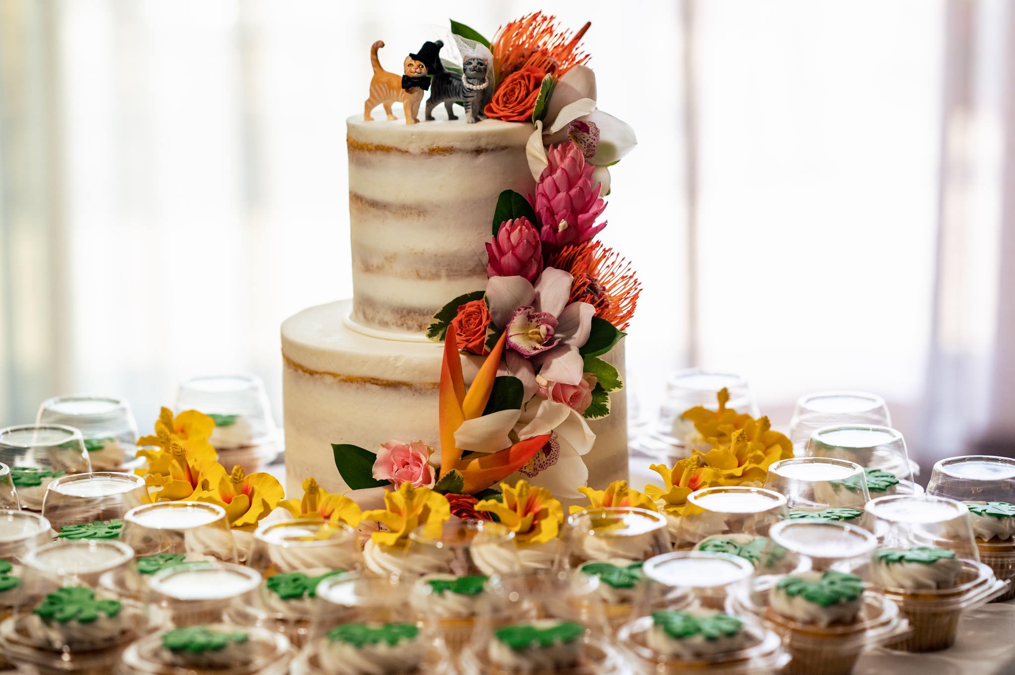 A two-tiered wedding cake adorned with colorful flowers and a cake topper featuring two small cat figurines, surrounded by individual dessert cups, perfect for a destination wedding in Honolulu.