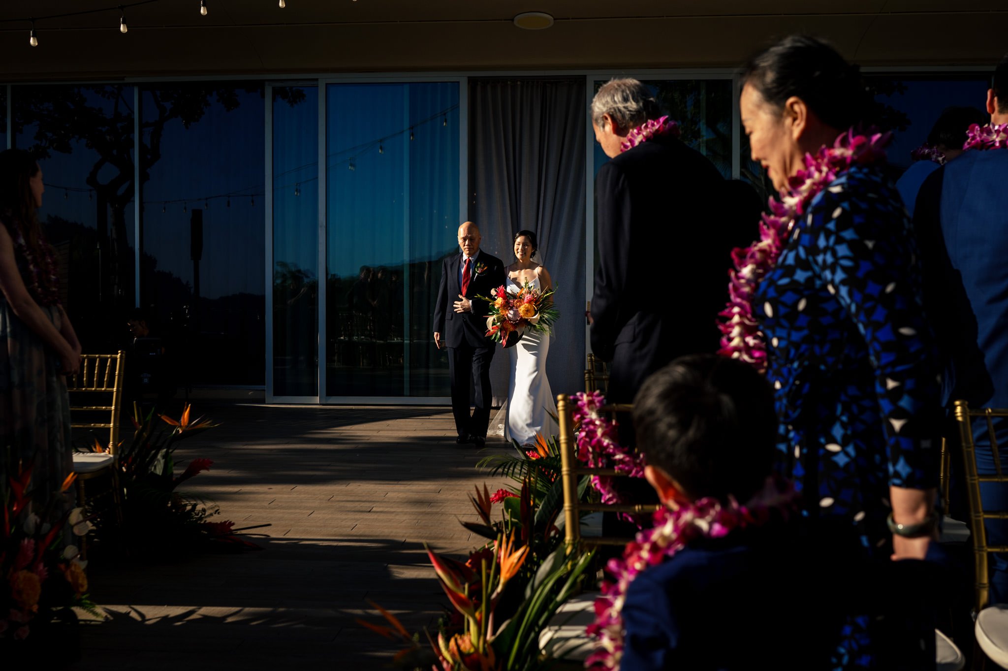 Bride walking down the aisle with her father, both smiling, with guests in Hawaiian leis watching at a sunny destination wedding in Honolulu.