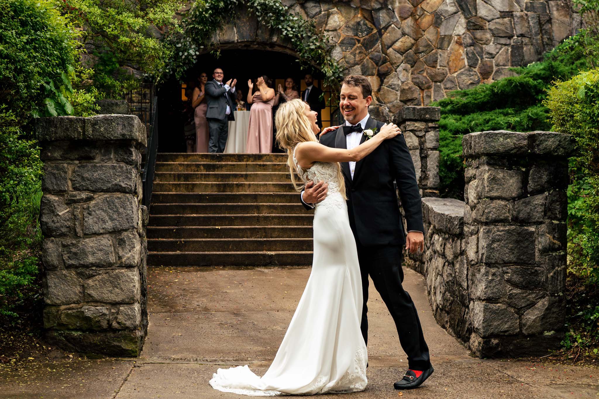 Bride and groom dancing joyfully outdoors at a Homewood wedding in Asheville, NC, with guests watching from a stone archway in a lush garden setting.