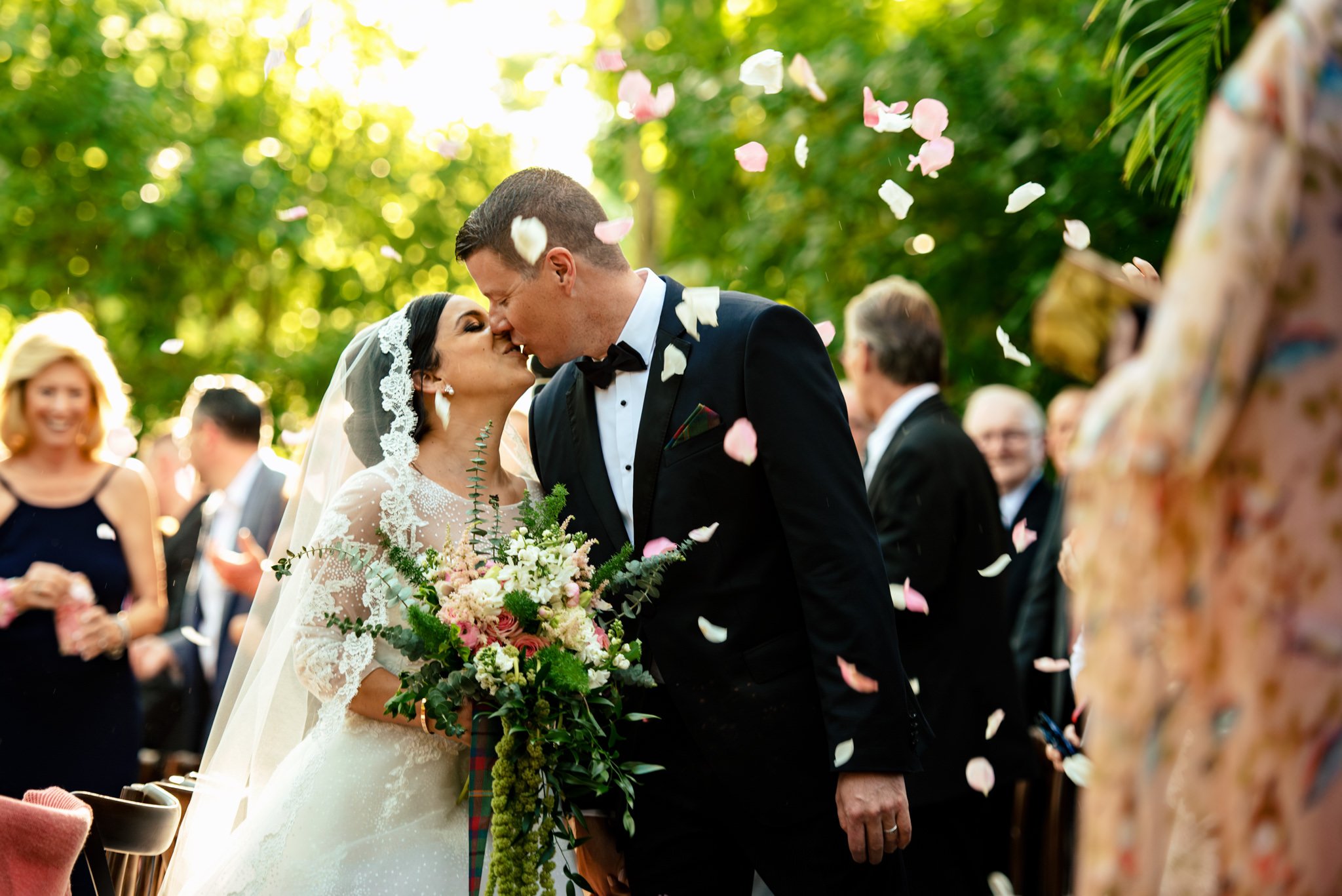 A bride and groom kiss under falling flower petals while walking down the aisle during an outdoor ceremony. Captured by Asheville, NC wedding photographer Michael Freas, guests are visible in the background.