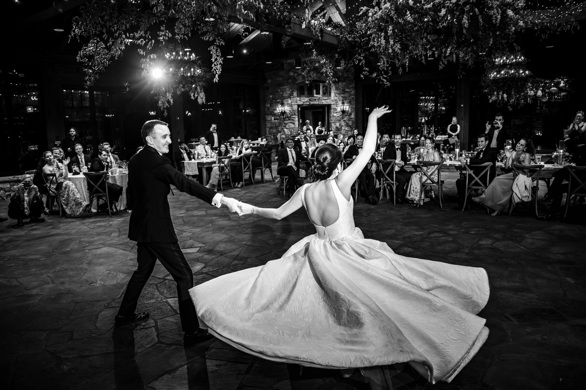 A couple in formal attire performs a dance in the middle of a reception hall with guests seated and watching in the background. Captured in black and white, this moment embodies the elegance of documentary wedding photography.