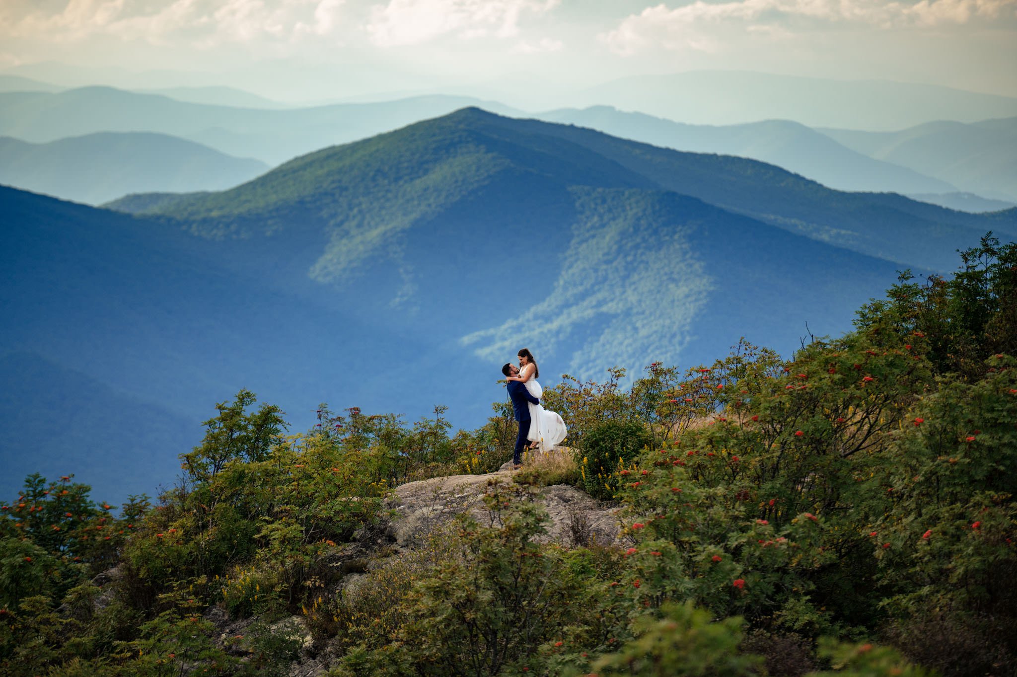 A couple embraces on a mountain peak, surrounded by lush greenery and layered mountains under a cloudy sky, celebrating their mountain elopement at Pretty Place Chapel.