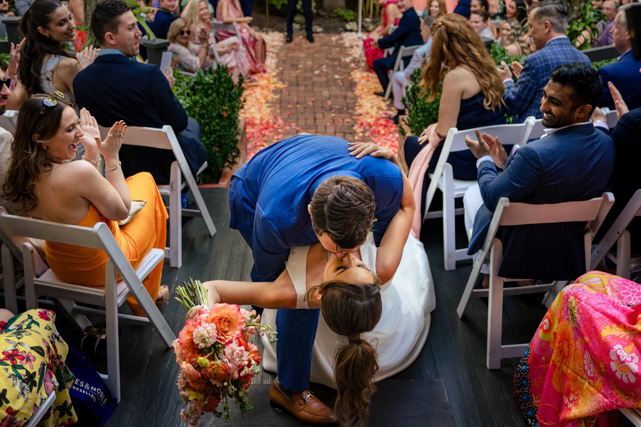 A bride and groom share a kiss at the end of the aisle while wedding guests applaud and smile. Captured by an Asheville Wedding Photographer, the joyful moment is made even more beautiful as the bride holds her bouquet of flowers.