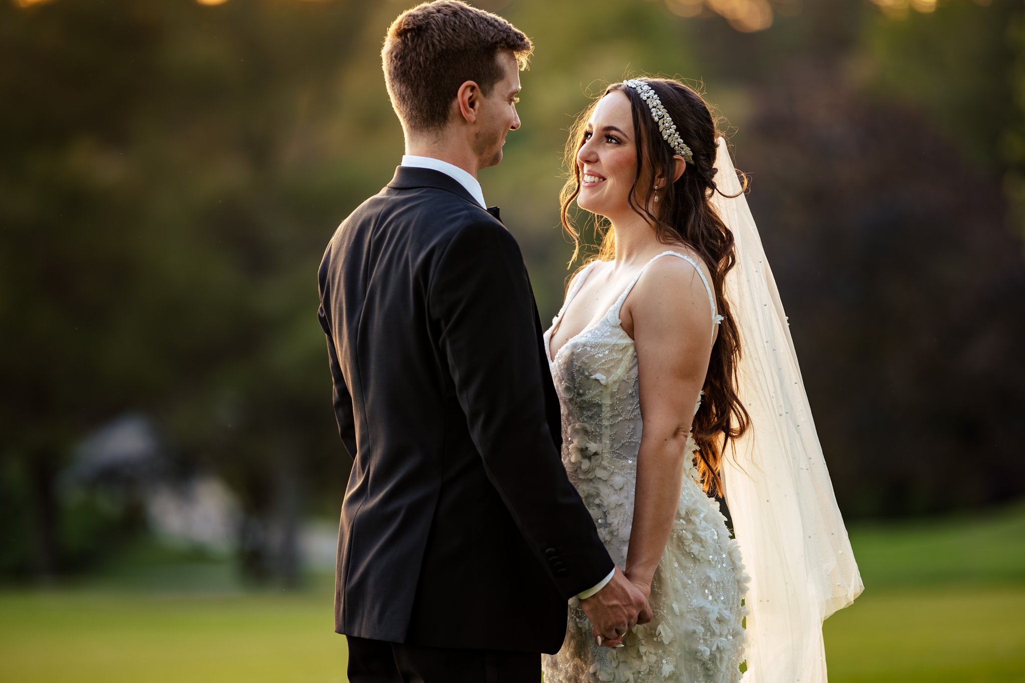 A bride and groom stand outdoors, facing each other and holding hands in wedding attire with greenery behind them—a perfect moment to consider when exploring photography packages for your special day.