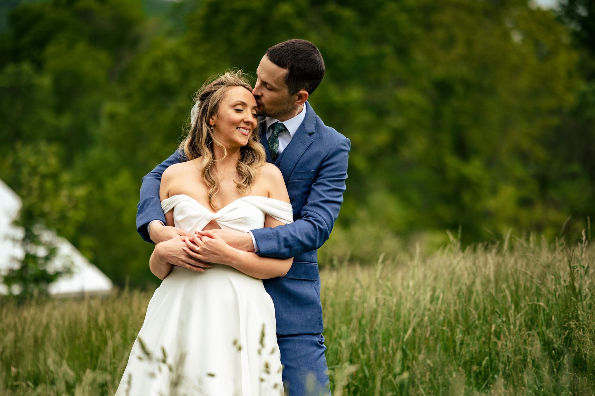 A bride and groom stand in a grassy field. The groom embraces the bride from behind as they both smile, surrounded by greenery—a perfect moment captured for photography guides or to inspire your next wedding photography pricing guide.
