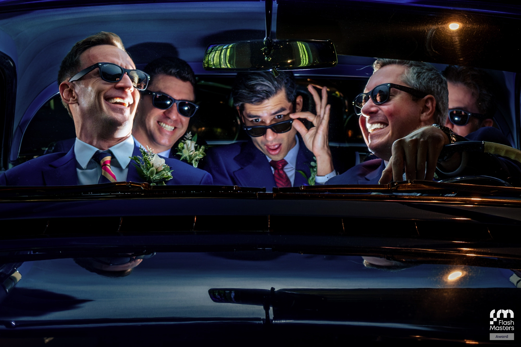 Five men in suits and sunglasses sit inside a car at night, smiling and laughing, with one man adjusting his glasses and another holding the steering wheel—captured perfectly by Flash Wedding Photography.