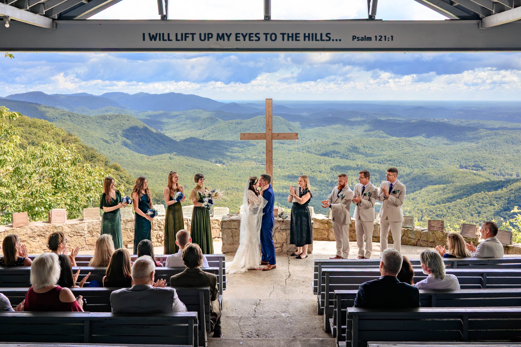 A couple stands at the altar with their wedding party in an outdoor chapel overlooking mountains—a perfect scene for Pretty Place weddings—where a wooden cross and seated guests complete the breathtaking setting.