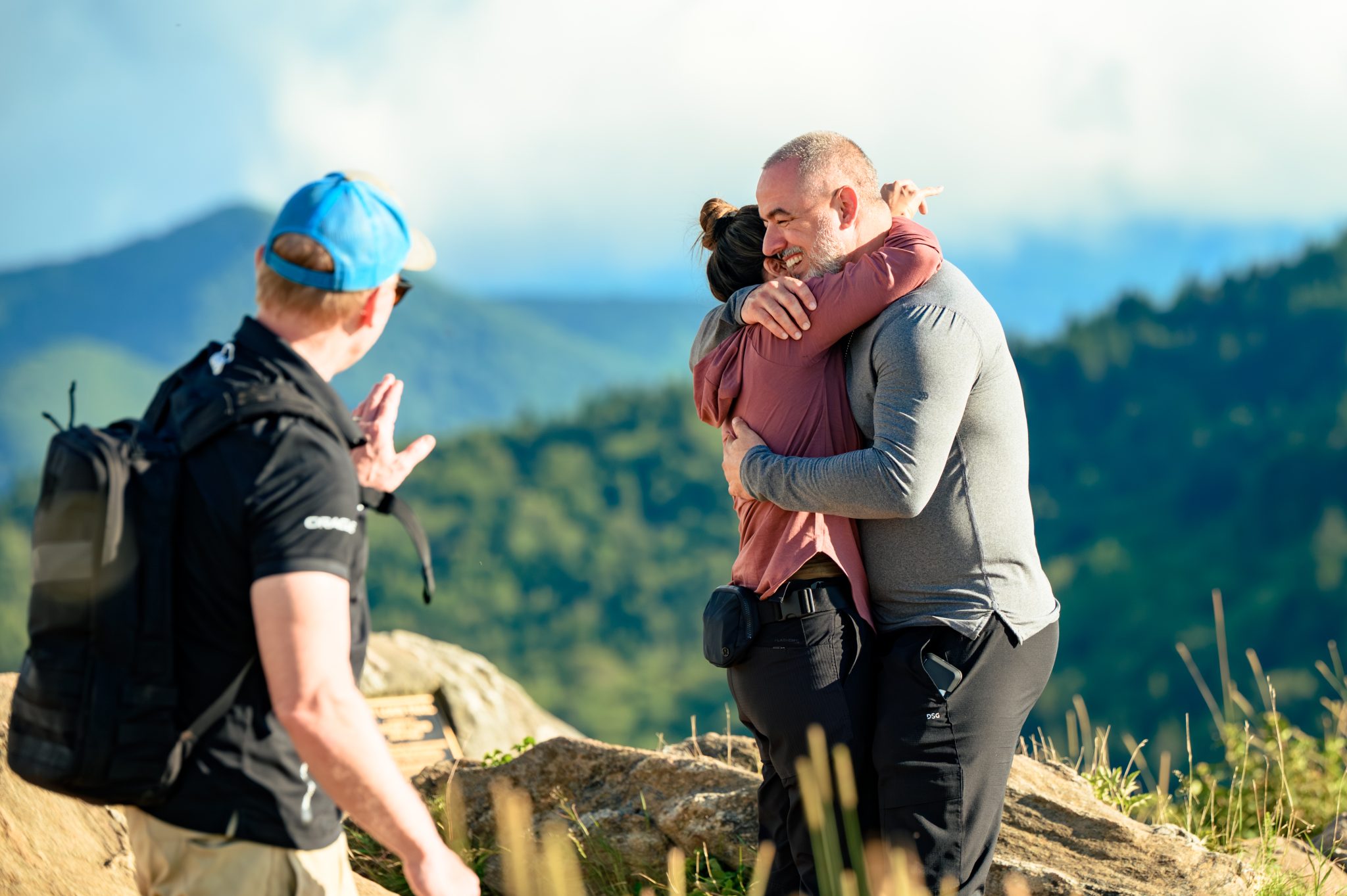 A man and woman embrace on a mountain overlook while another man with a backpack waves at them, creating a moment worth sharing in testimonials. The background features green hills and a cloudy sky.