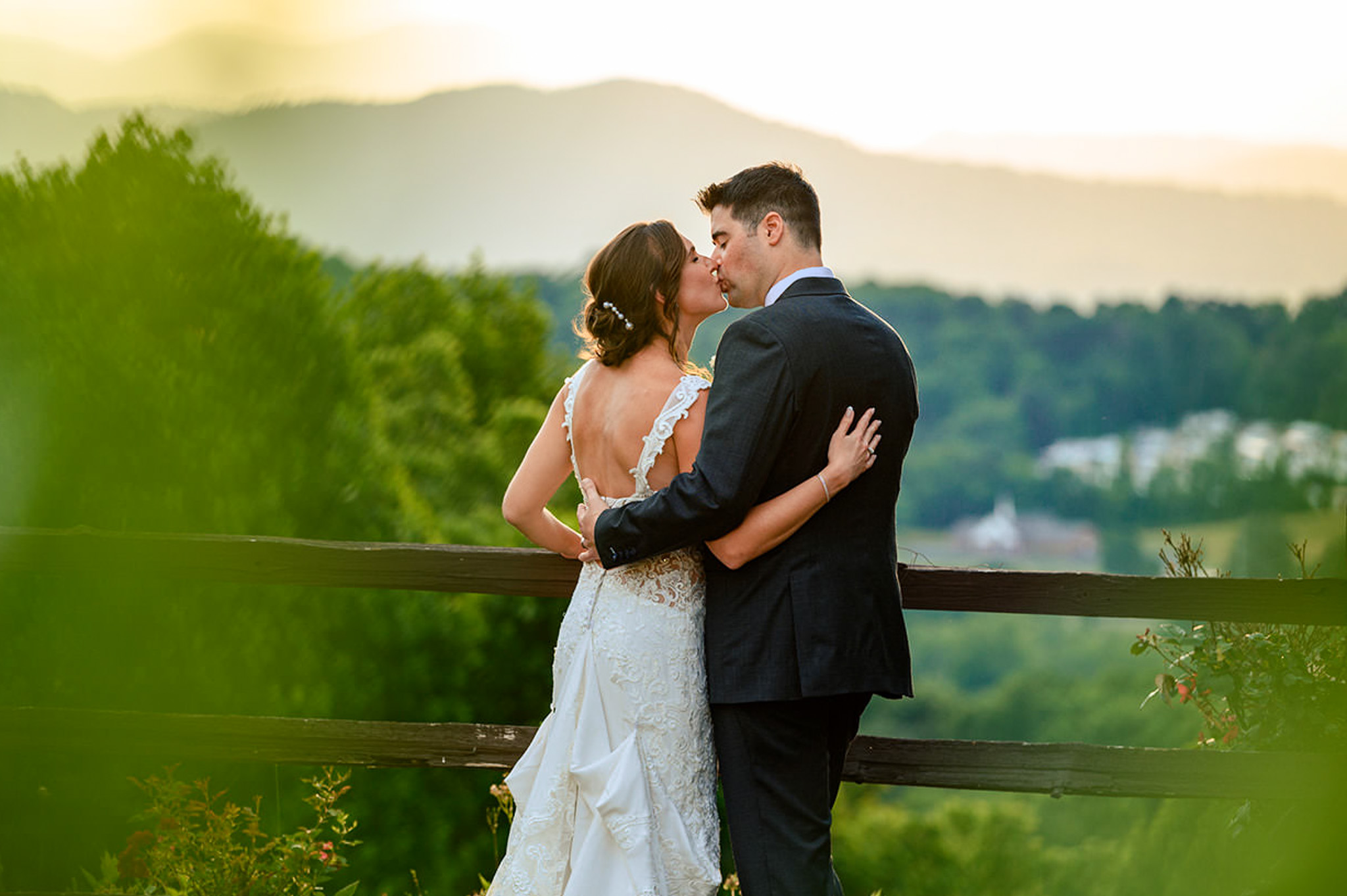 A bride and groom embrace and kiss outdoors by a wooden fence, surrounded by lush greenery and distant Blue Ridge Mountains at sunset—captured beautifully by a Blue Ridge Mountains wedding photographer.