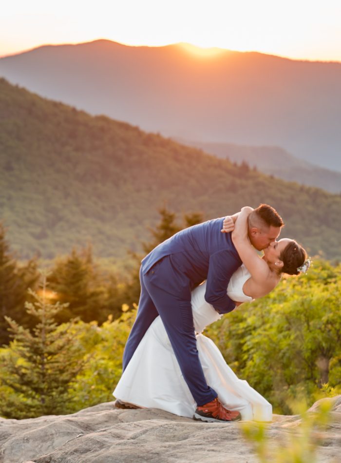 A bride and groom share a kiss during a dip on a rocky overlook with green mountains and a setting sun in the background, captured perfectly by a documentary wedding photographer.