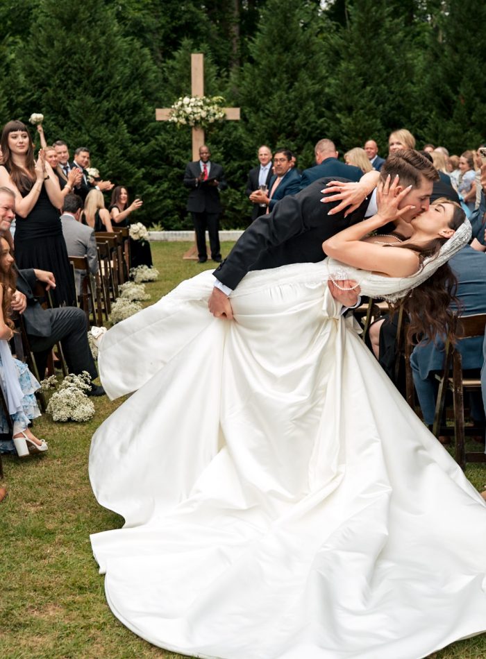 A groom dips and kisses his bride in the aisle after their outdoor wedding ceremony, as guests stand, smile, and clap—captured perfectly by a documentary wedding photographer.