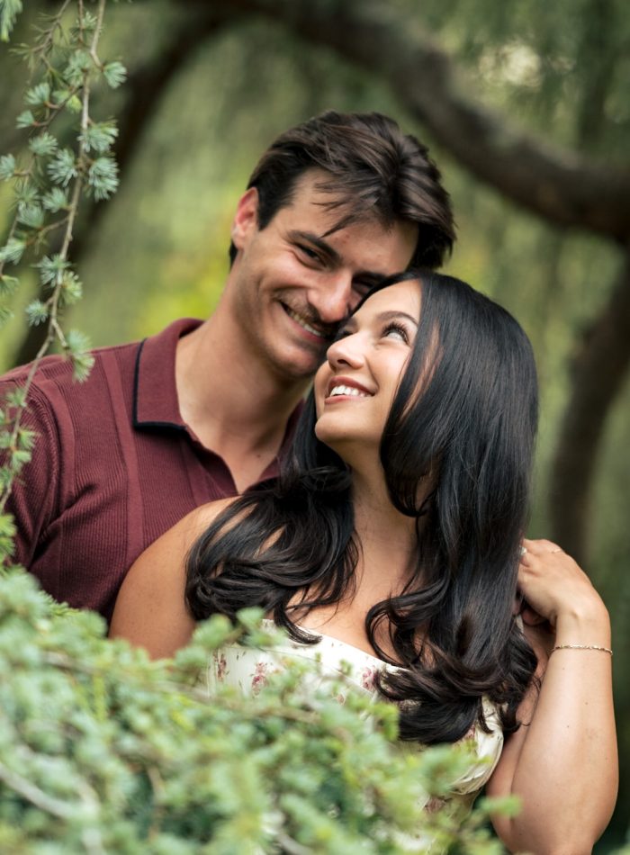 A man and woman stand close together, smiling and looking at each other, surrounded by greenery in an outdoor setting, beautifully captured by a documentary wedding photographer.