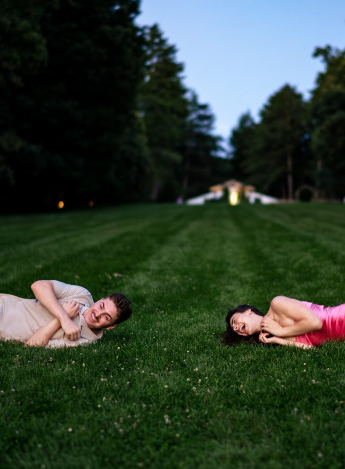 Two people rolling down the Diana hill aat Biltmore Estate in Asheville NC, both resting on their sides. One wears jeans and a shirt, the other a pink dress. Trees and a distant structure are in the background.
