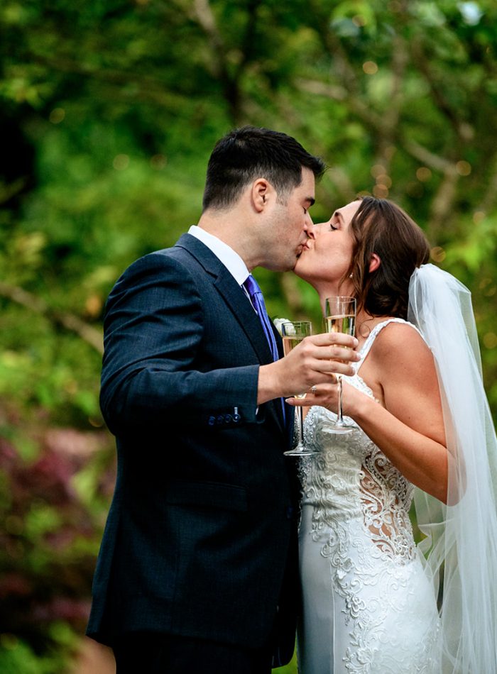 A bride and groom in formal attire kiss outdoors while holding champagne glasses, surrounded by greenery—an unforgettable moment captured beautifully by an Asheville wedding photographer.