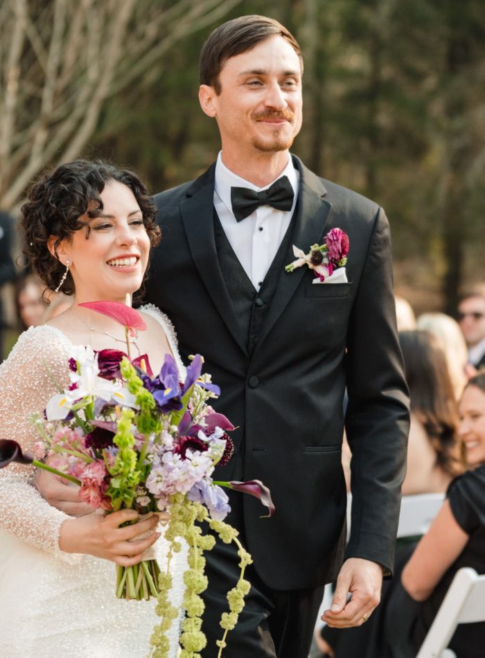 A bride in a white dress holds a bouquet and stands next to a groom in a black tuxedo; guests are seated outdoors in the background.