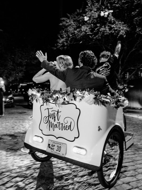 A couple rides away in a decorated pedicab with a "Just Married" sign at night, waving as they leave a cobblestone street—capturing the perfect ending to their special day and highlighting the value of thoughtful wedding photography pricing.