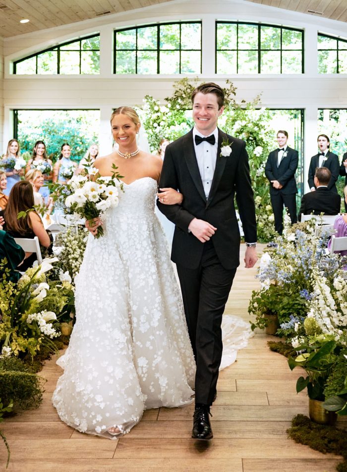 A bride and groom walk down the aisle together after their wedding ceremony in a bright indoor venue, surrounded by guests and floral arrangements—a perfect moment to remember when exploring wedding photography pricing for your special day.