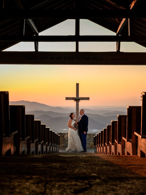 A bride and groom stand in front of a cross at an outdoor chapel overlooking mountains at sunset, beautifully captured by an Asheville elopement photographer.