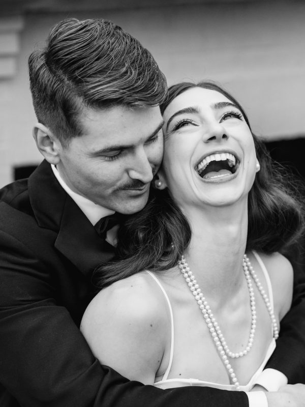 A man in a suit hugs a smiling woman wearing a pearl necklace and a white dress. Both appear joyful, with the woman laughing heartily—a perfect candid moment often treasured in wedding photography pricing collections.